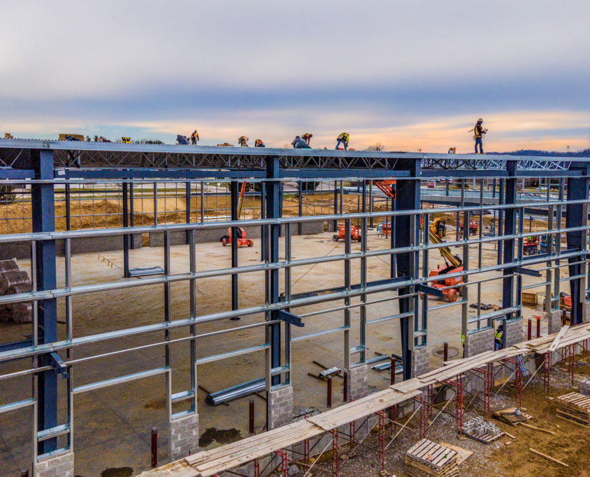 A crew installing metal roof decking along the top of a steel-framed commercial building at dusk, with masonry walls and boom lifts on the active site below