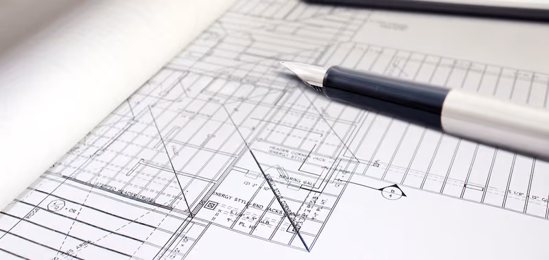 A close-up of a printed floor plan — dimension strings, wall types, and framing callouts — with a draftsman’s pen resting across the sheet