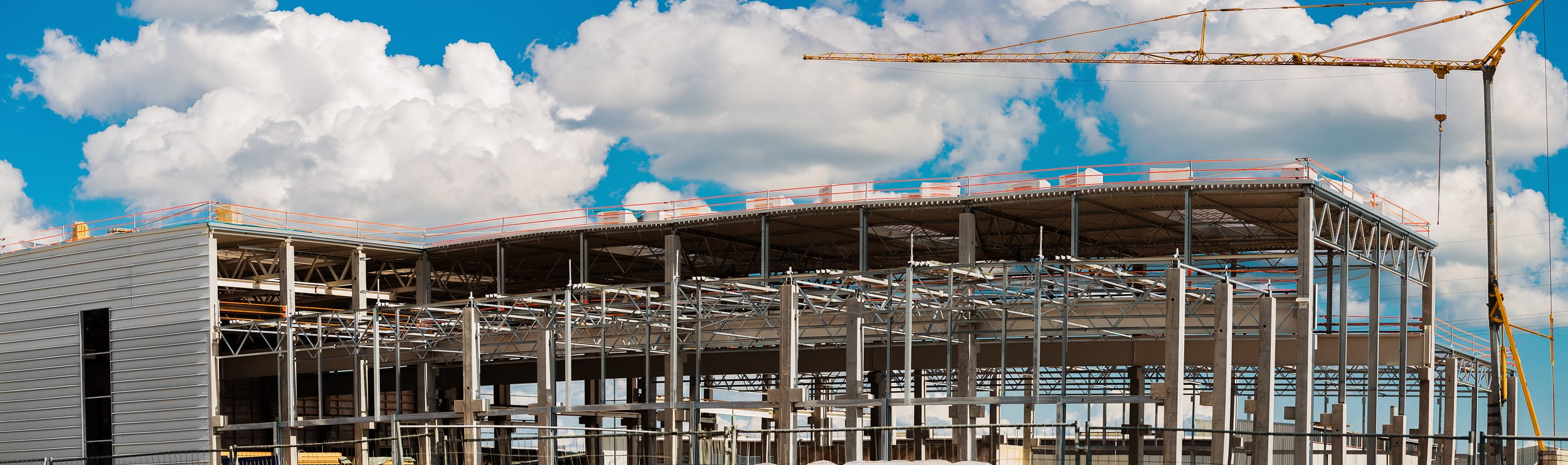 A steel-framed commercial building under construction against a bright cumulus sky, with metal roof decking in place, edge-protection railing along the perimeter, and a tower crane jib reaching across the site
