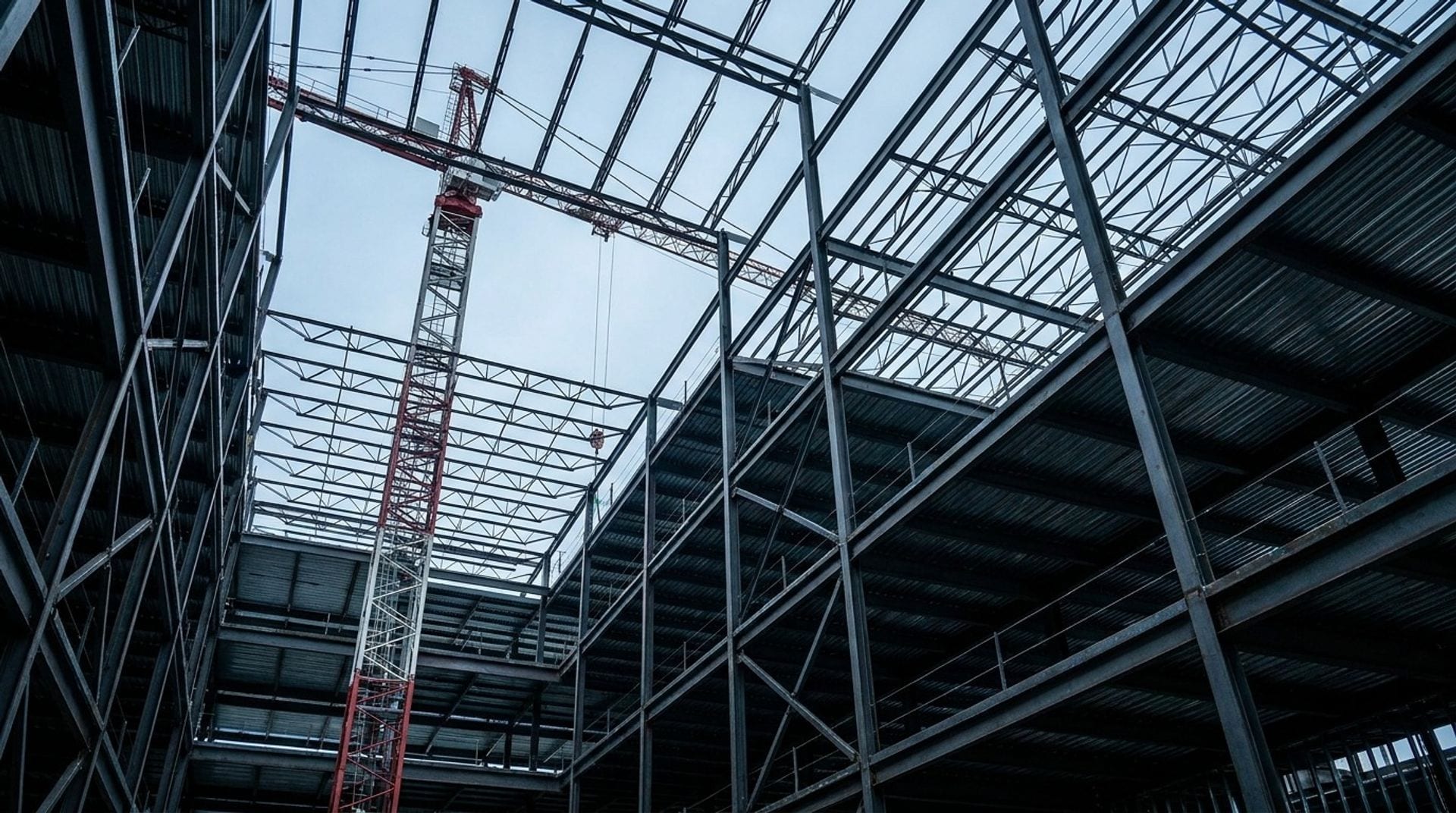 A tower crane rising through the dense structural steel framing of a large commercial building under construction, seen from below against an overcast sky