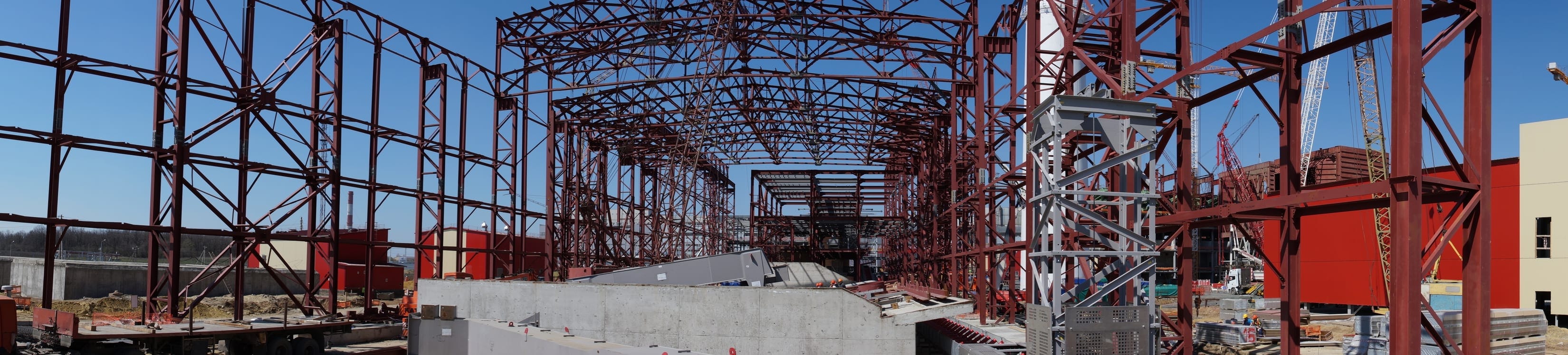 Red structural steel framing rising across a large industrial job site, with roof trusses spanning overhead, crawler cranes at work, and poured concrete walls in the foreground