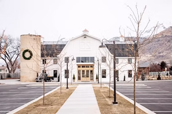 The finished Walker Farms Event Center — a white barn-style venue with a restored silo and gabled entry, fronted by a new landscaped walkway and parking lot below the Wasatch foothills