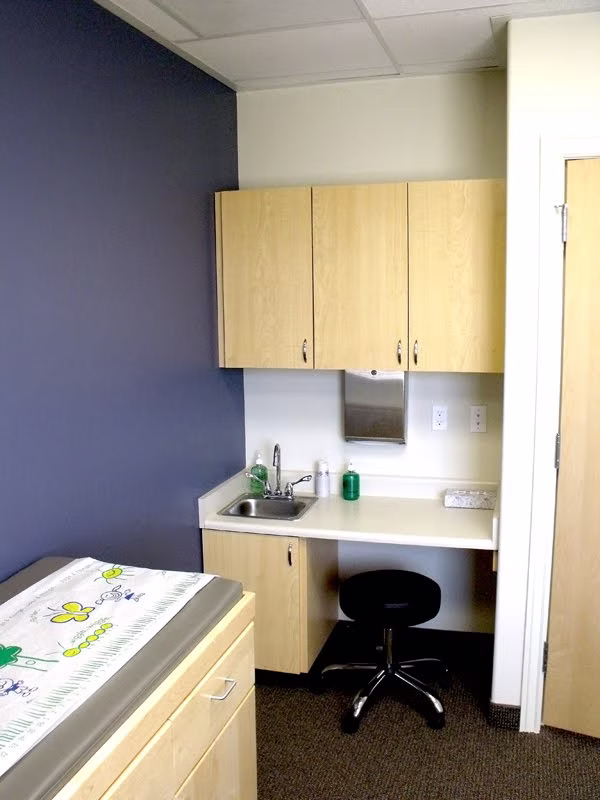 A completed exam room at Utah Valley Pediatrics — maple casework and a sink counter against a deep blue accent wall, with a papered pediatric exam table alongside