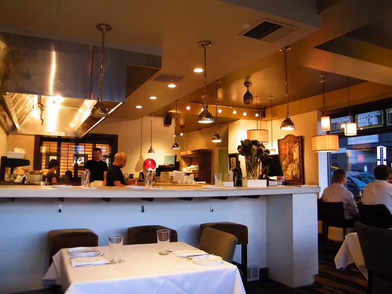 The finished Communal dining room — white-linen tables in front of a long service bar, lit by a row of pendant lamps with the open kitchen beyond